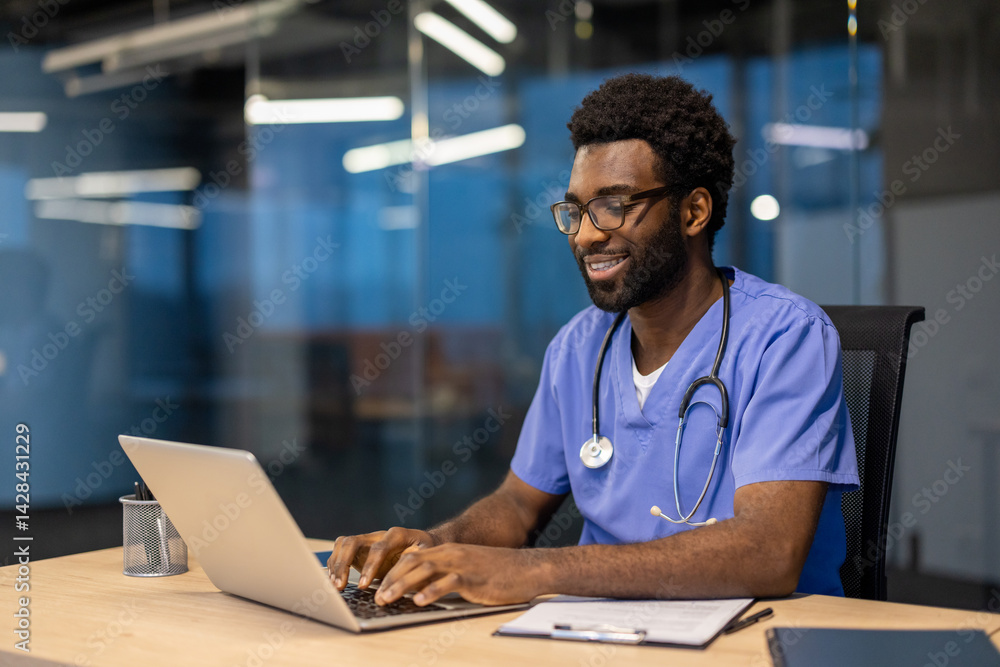 A smiling doctor in scrubs works on a laptop, seated at a desk in a modern office. A stethoscope hangs around his neck.