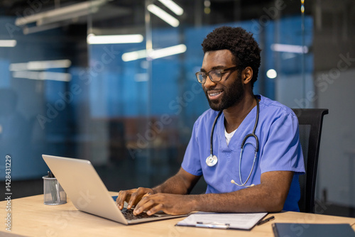Obraz na płótnie A smiling doctor in scrubs works on a laptop, seated at a desk in a modern office. A stethoscope hangs around his neck. w salonie
