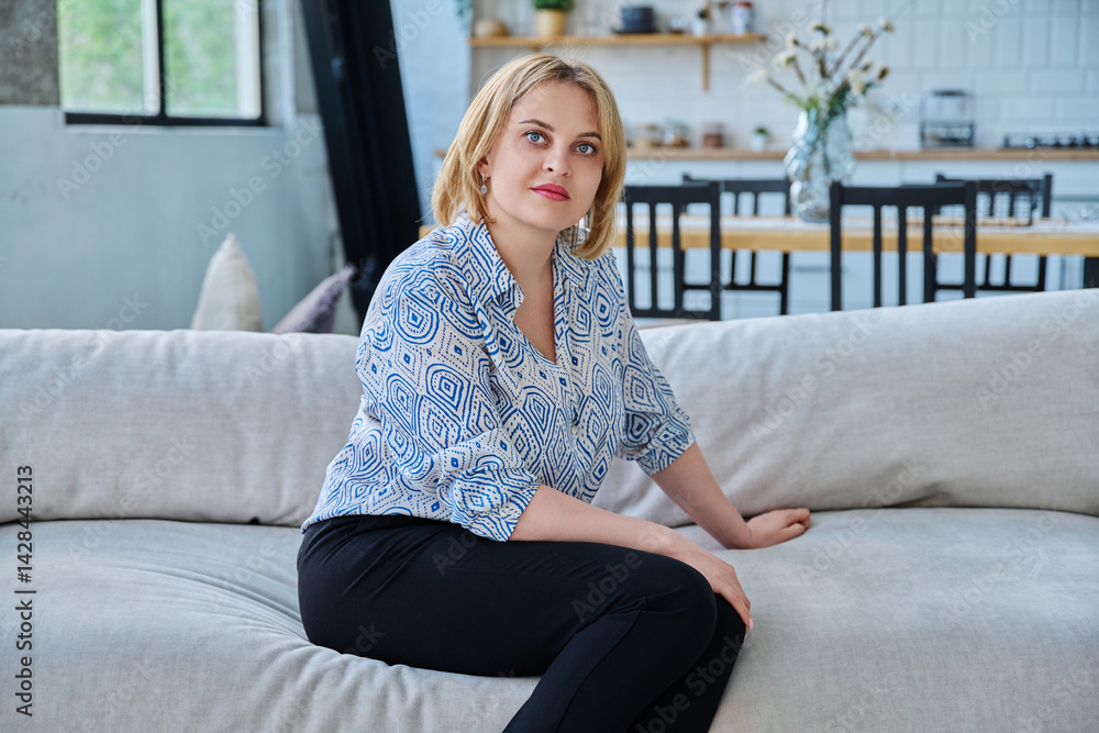 Portrait of middle aged blonde woman with blue eyes sitting on the sofa in the living room.