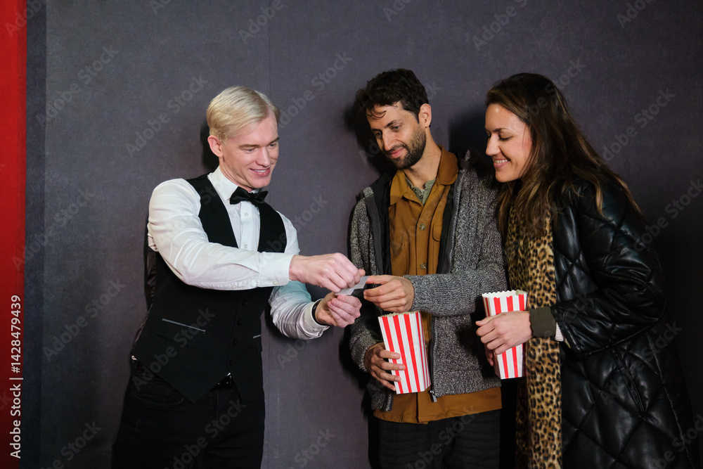 Usher tearing movie tickets of young couple holding popcorn buckets at cinema entrance