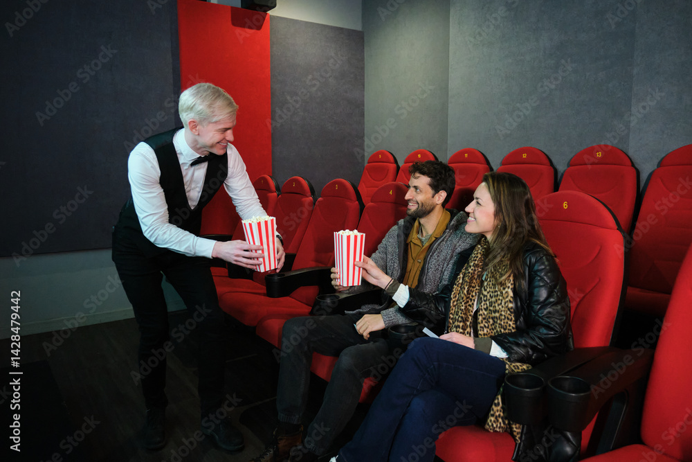 Waiter bringing popcorn buckets to young couple sitting in movie theater