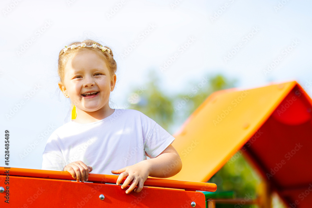 Happy blonde girl in a white T-shirt with a floral headband smiling joyfully on a vibrant, colorful playground. The background is full of playful red, yellow, green, and blue elements.