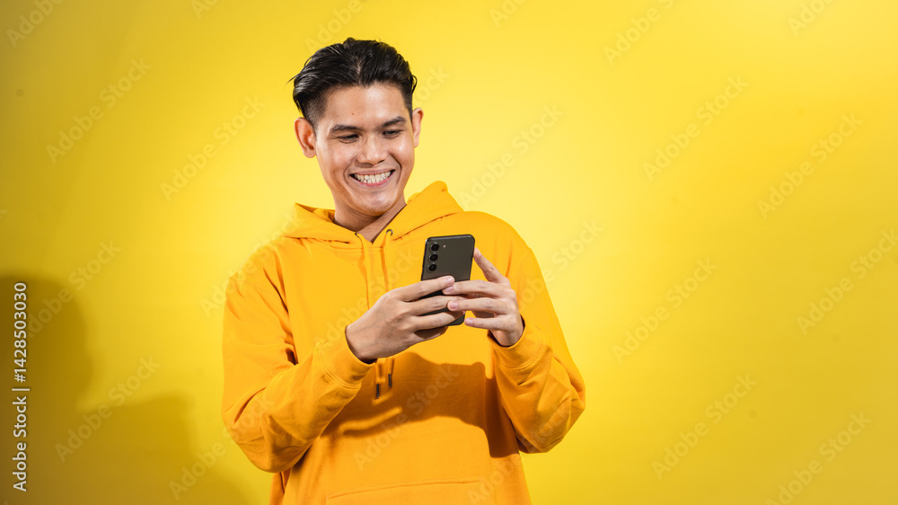 Young Asian man in yellow hoodie smiling while using smartphone against vibrant yellow background. Studio portrait showing digital lifestyle and modern communication in cheerful tone Young Asian man in yellow hoodie smiling while using smartphone against vibrant yellow background. Studio portrait showing digital lifestyle and modern communication in cheerful tone