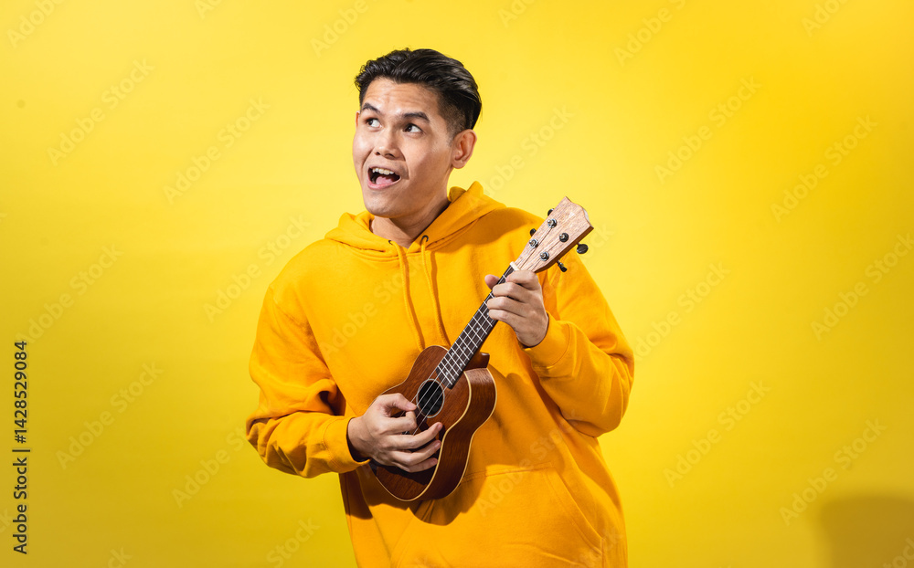 Asian man in yellow hoodie enthusiastically playing a small ukulele against a vivid yellow background. Expressive musical portrait with happy mood and creative, artistic personality