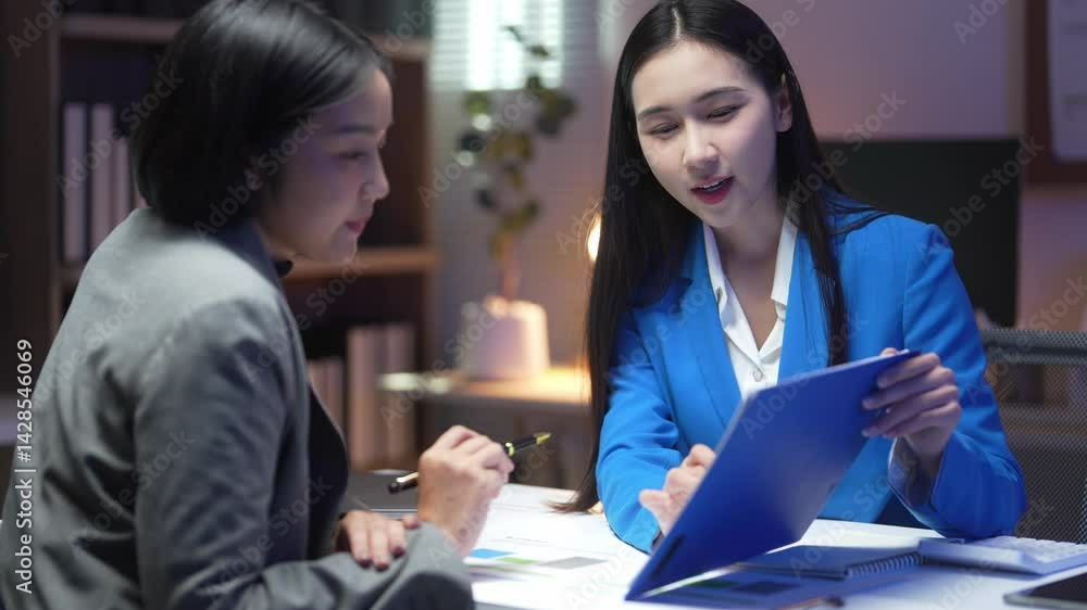 Two women are sitting at a table, one of them holding a blue folder. They are discussing something important, possibly related to work or a project. Scene is serious and focused Two women are sitting at a table, one of them holding a blue folder. They are discussing something important, possibly related to work or a project. Scene is serious and focused