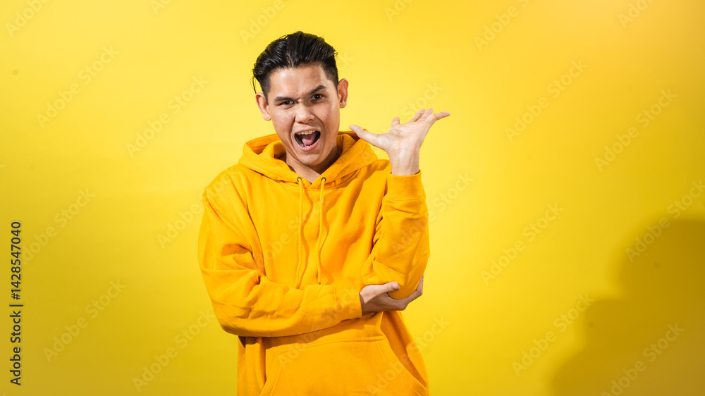 Playful young Asian man in yellow hoodie raising one hand with dramatic sassy expression and open mouth, posing against solid yellow background. Studio portrait for bold, expressive attitude Playful young Asian man in yellow hoodie raising one hand with dramatic sassy expression and open mouth, posing against solid yellow background. Studio portrait for bold, expressive attitude
