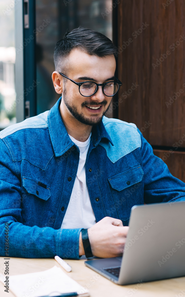 Happy young man in eyewear browsing netbook in summer area of urban cafeteria