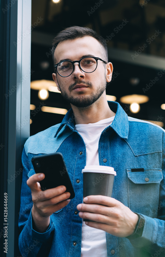 Serious bearded guy messaging on smartphone and holding tea in disposable cup in modern cafeteria