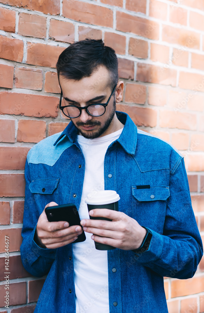 Focused male in eyeglasses messaging on smartphone while drinking tea against brick wall in street
