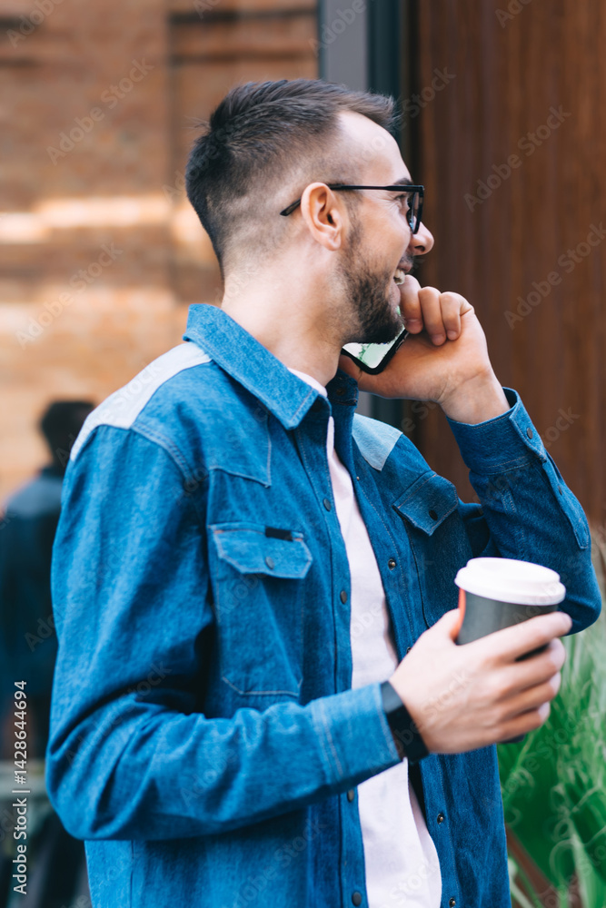 Happy male in glasses speaking on phone while strolling with takeaway coffee in street