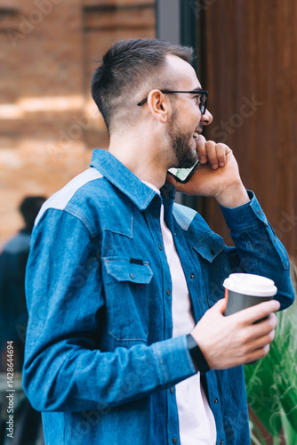 Obraz na płótnie Happy male in glasses speaking on phone while strolling with takeaway coffee in street w salonie