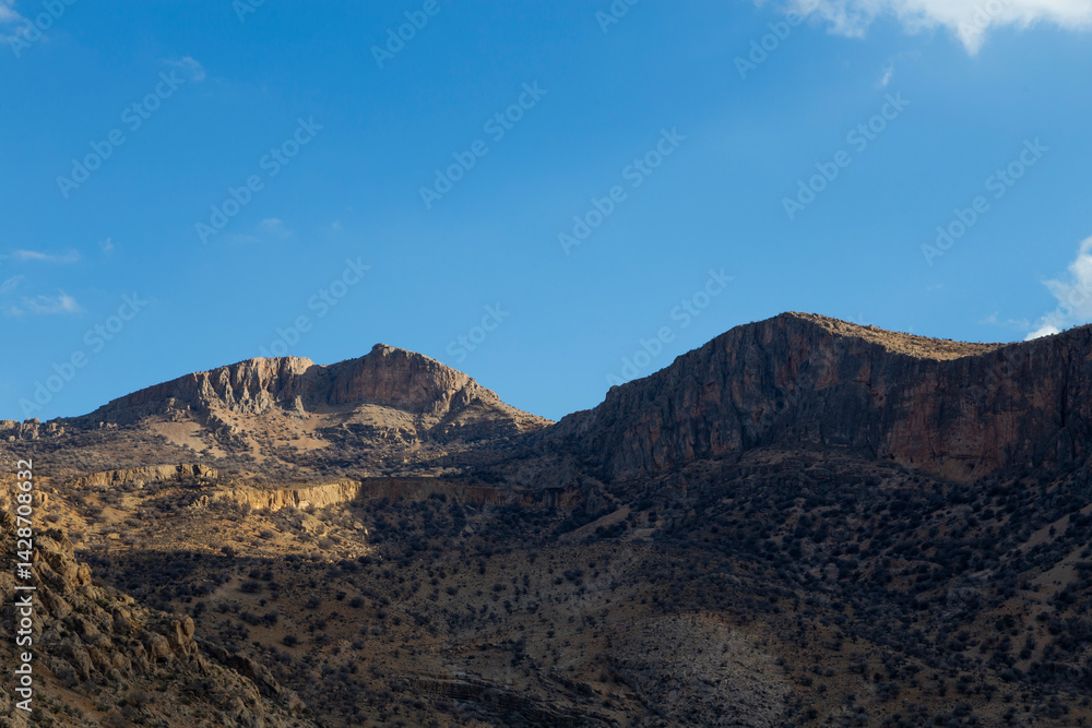 Rugged mountains bathed in warm sunlight, towering over rocky terrain with scattered vegetation, creating a timeless landscape of natural beauty in Fars province. Rugged mountains bathed in warm sunlight, towering over rocky terrain with scattered vegetation, creating a timeless landscape of natural beauty in Fars province.