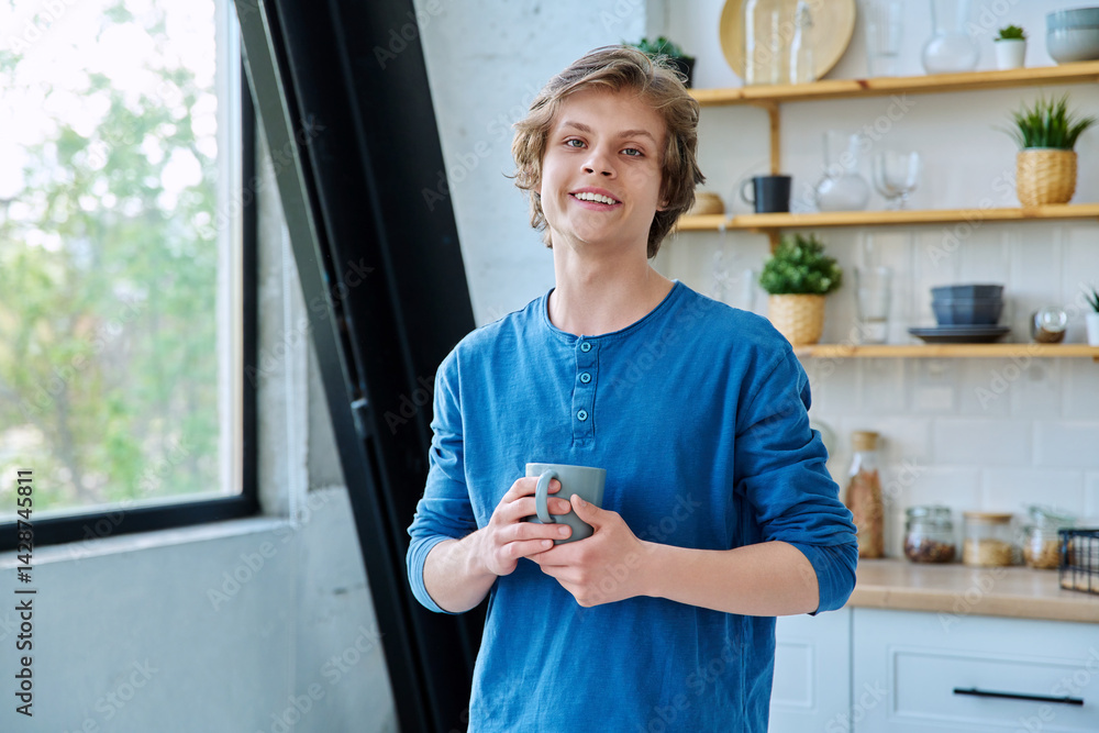 Teenage smiling guy with cup in hands in home interior, looking at camera
