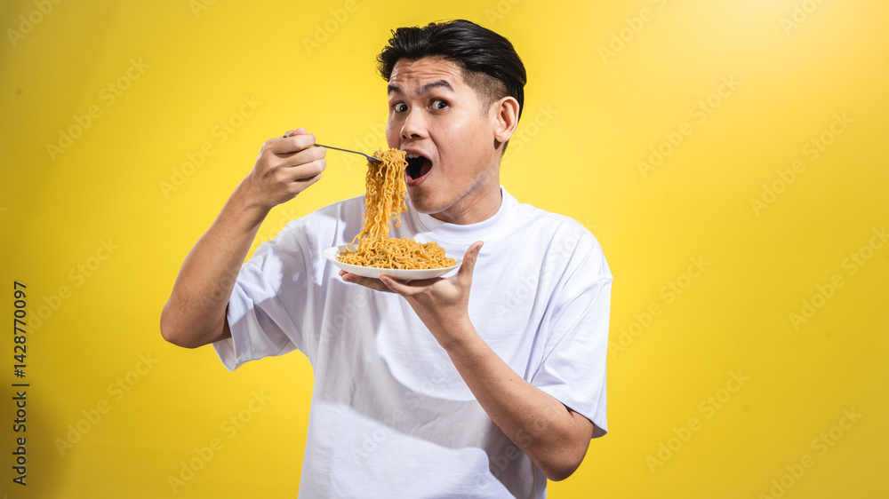 Asian man in a white t-shirt enthusiastically eating noodles from a plate with a fork, looking to the side with a playful expression, standing against a bright yellow background Asian man in a white t-shirt enthusiastically eating noodles from a plate with a fork, looking to the side with a playful expression, standing against a bright yellow background
