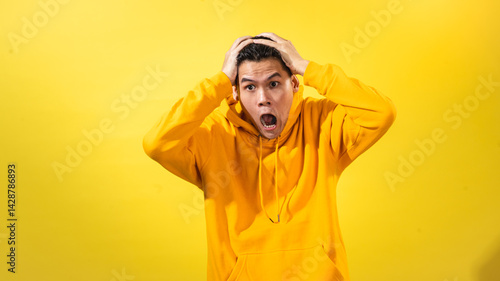 Shocked young Asian man in bright yellow hoodie holding head with both hands, mouth open wide in disbelief, posing against solid yellow background. Concept of panic, stress, or surprise