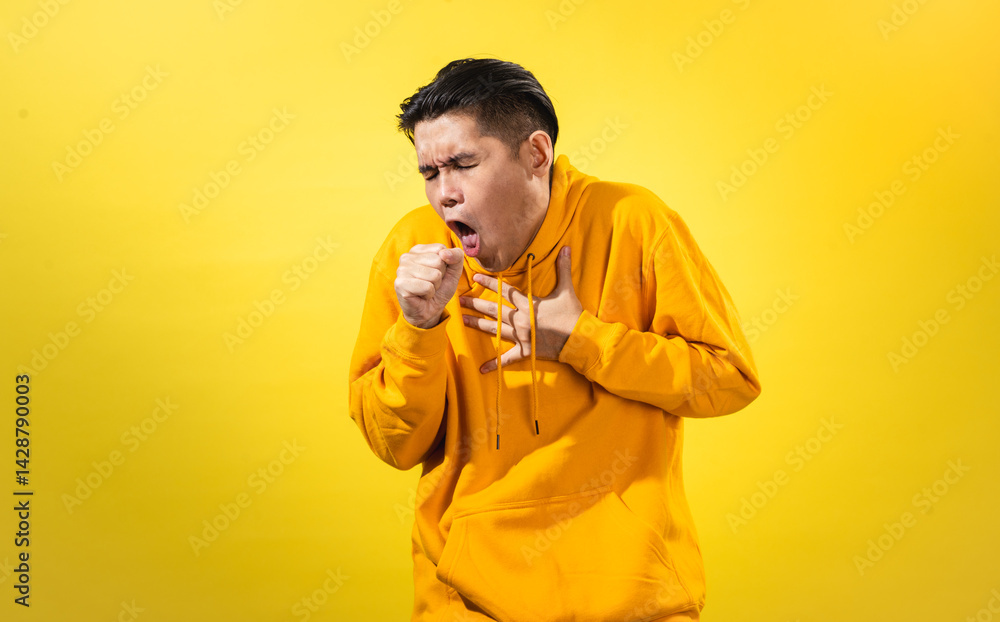 Unwell young Asian man in yellow hoodie coughing into his fist and holding his chest with discomfort, eyes closed, standing against solid yellow background. Studio concept for cold, flu, or sickness