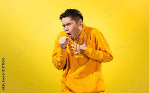 Obraz na płótnie Unwell young Asian man in yellow hoodie coughing into his fist and holding his chest with discomfort, eyes closed, standing against solid yellow background. Studio concept for cold, flu, or sickness w salonie