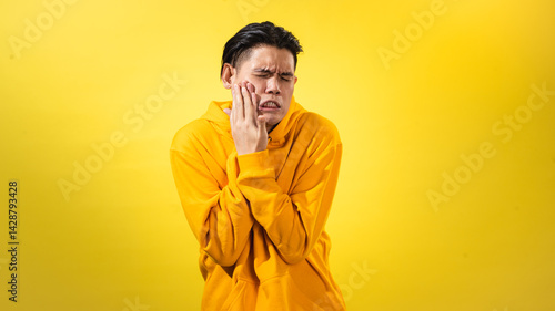 Asian man in yellow hoodie pressing his cheek in pain, eyes shut tight, showing discomfort from toothache or jaw pain, against solid yellow background. Studio shot expressing oral health issue