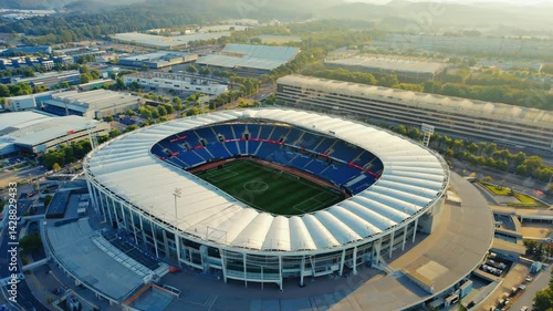 Aerial view of a large open air stadium with blue seating and green playing field and surrounding cityscape in the daytime.