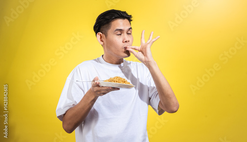 Asian man in a white t-shirt tasting fried noodles with a satisfied expression, eyes closed, and making a 