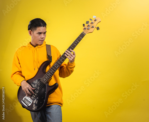 Asian man in a yellow hoodie playing an electric bass guitar against a bright yellow background. Captured mid-performance with a focused expression and casual modern style