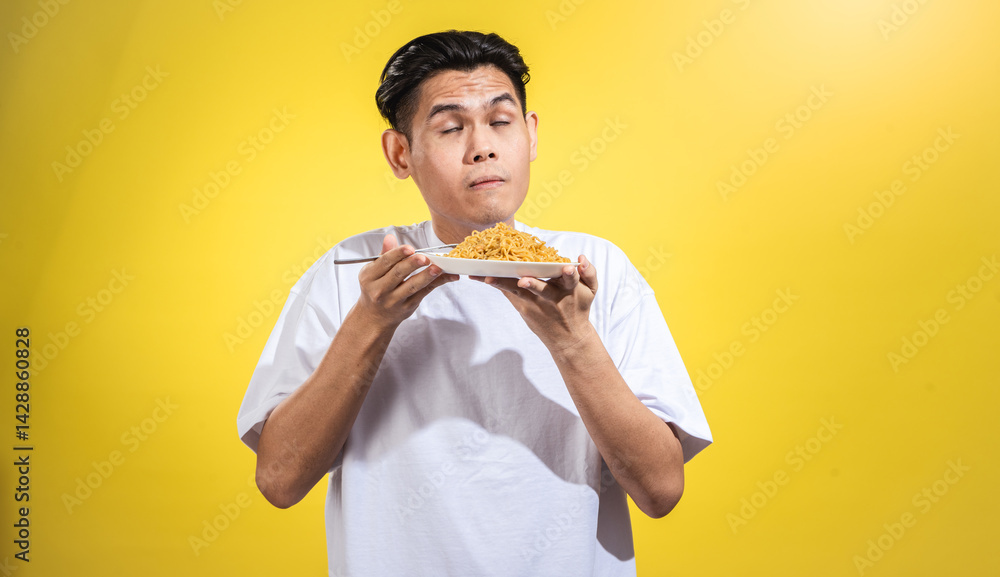 Asian man in white t-shirt holding a plate of noodles near his face with closed eyes and a satisfied expression, as if savoring the aroma, against a bright yellow background Asian man in white t-shirt holding a plate of noodles near his face with closed eyes and a satisfied expression, as if savoring the aroma, against a bright yellow background