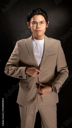 Asian man with glasses wearing beige suit and white shirt poses confidently with hands joined in front of chest against dark studio background, ideal for corporate or branding themes
