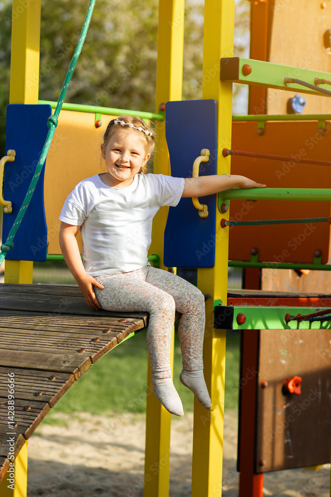 Happy blonde girl in a white T-shirt with a floral headband smiling joyfully on a vibrant, colorful playground. The background is full of playful red, yellow, green, and blue elements.