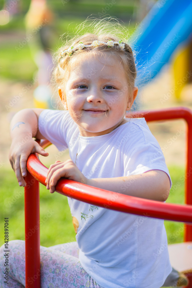 Fototapeta premium Happy blonde girl in a white T-shirt with a floral headband smiling joyfully on a vibrant, colorful playground. The background is full of playful red, yellow, green, and blue elements. 