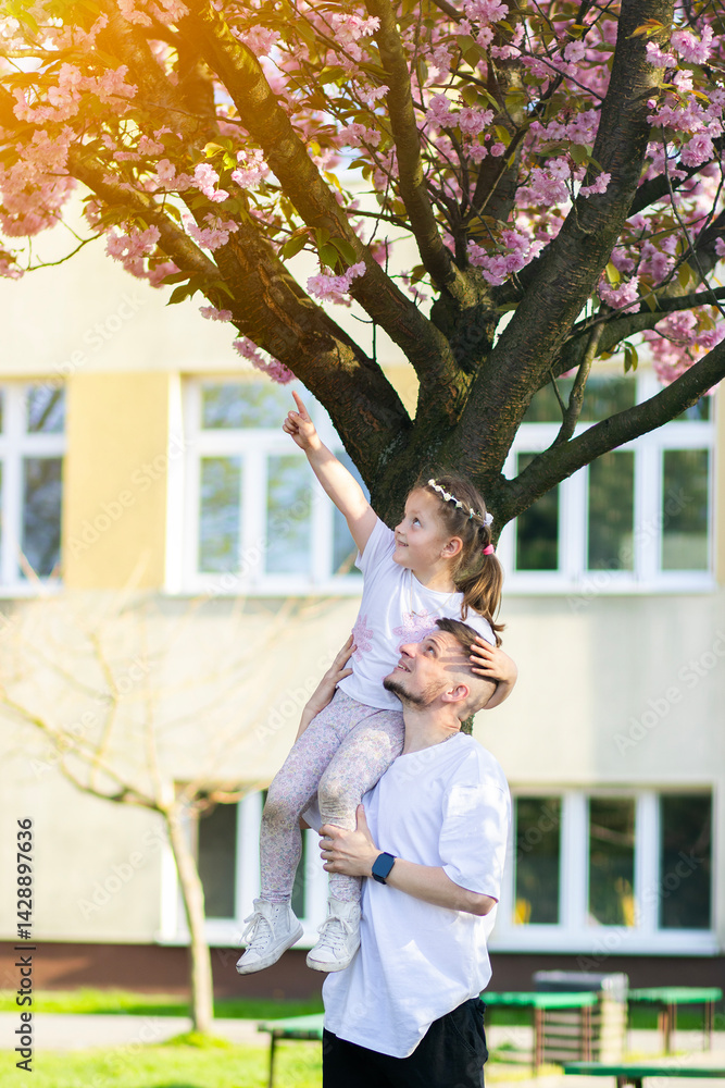 Man in white T-shirt and black pants holding a little girl in white shirt, pointing at blossoming tree. Springtime scene with bright flowers and greenery in the background.