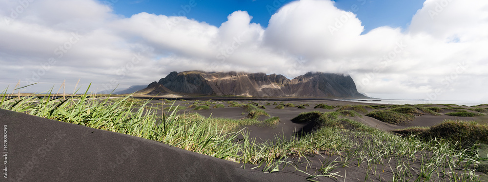 Low-angle panorama of windswept black sand dunes in front of rugged Icelandic mountains partially covered by dramatic storm clouds.
