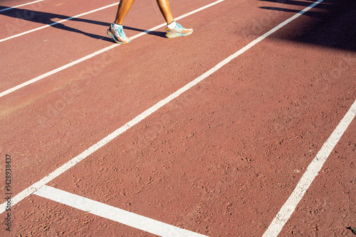 A close-up of a runner's legs walking on an outdoor athletics track with clear white lane markings under bright sunlight, illustrating sports and healthy lifestyle concepts.