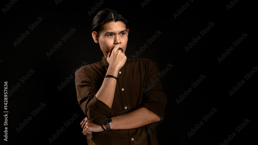 Asian man in dark brown shirt standing in studio with dark background, touching lips with thoughtful expression, perfect for concept visuals related to thinking, planning, or personal reflection