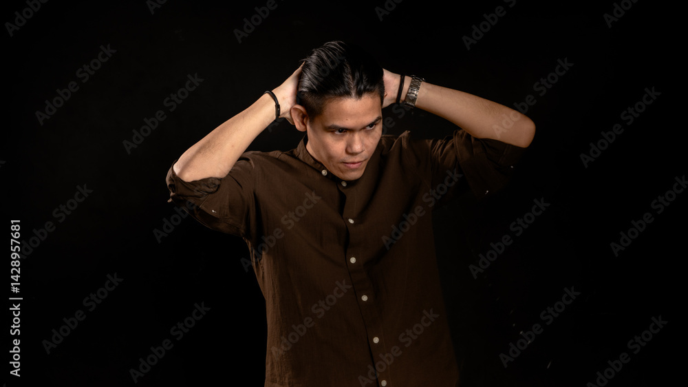 Asian man in dark brown shirt styling his hair with both hands behind head in studio with dark background, showing confidence and focus, ideal for fashion, grooming, or personal branding visuals