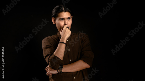 Obraz na płótnie Asian man in dark brown shirt standing in studio with dark background, touching lips with thoughtful expression, perfect for concept visuals related to thinking, planning, or personal reflection w salonie