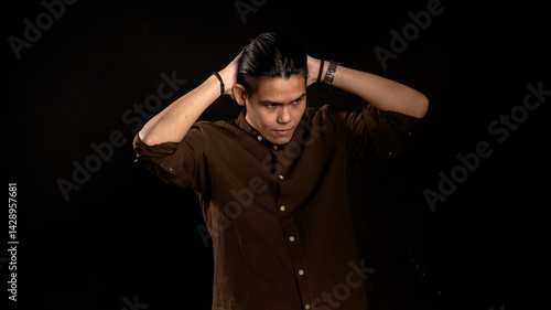 Obraz na płótnie Asian man in dark brown shirt styling his hair with both hands behind head in studio with dark background, showing confidence and focus, ideal for fashion, grooming, or personal branding visuals w salonie