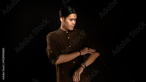 Obraz na płótnie Asian man in dark brown shirt rolling up sleeves while looking down with composed expression against black background, perfect for concepts of focus, preparation, productivity, or personal style w salonie