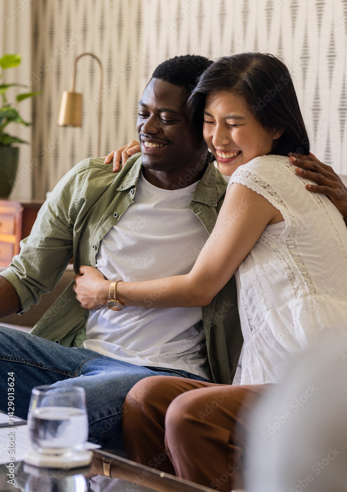 Diverse couple hugging on sofa in cozy living room, with wooden coffee table and potted plant Diverse couple hugging on sofa in cozy living room, with wooden coffee table and potted plant