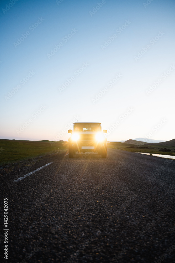 SUV with headlights on driving toward camera at sunset on an empty rural road, with mountains and grass fields in the distance.