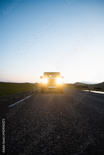 Obraz na płótnie SUV with headlights on driving toward camera at sunset on an empty rural road, with mountains and grass fields in the distance. w salonie
