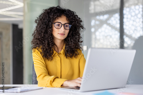 Fotografie A woman with curly hair, wearing glasses and a yellow shirt, works on a laptop in an office setting