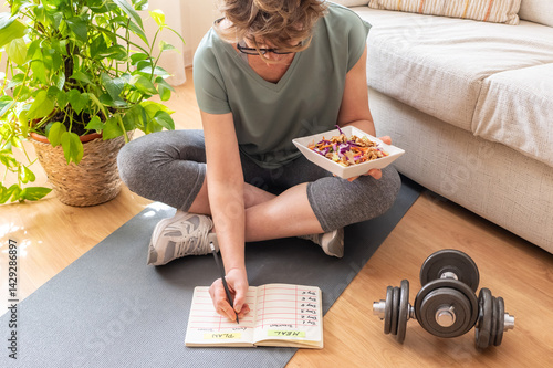 Sitting on a yoga mat, a mature woman writes down her meal plan while holding a healthy salad, with dumbbells nearby, symbolizing her commitment to a balanced lifestyle