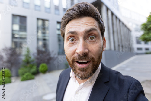 A surprised businessman looks directly at the camera outdoors with his mouth open in shock, in front of a modern building.