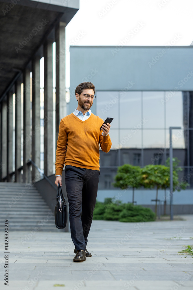 A stylish businessman walks outside, engrossed in his smartphone, carrying a briefcase near a modern building.