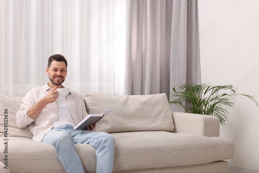 Happy man drinking coffee while reading book on sofa near window with beautiful curtains at home in living room. Space for text Happy man drinking coffee while reading book on sofa near window with beautiful curtains at home in living room. Space for text