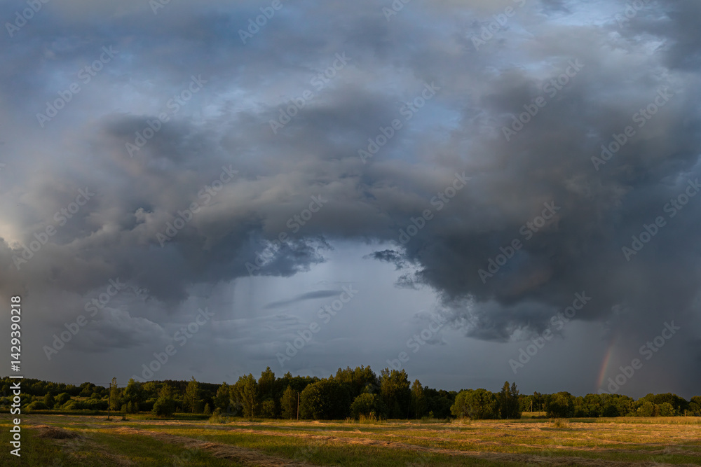 Faint Rainbow Beneath Storm Clouds Over Sun-Kissed Countryside Faint Rainbow Beneath Storm Clouds Over Sun-Kissed Countryside