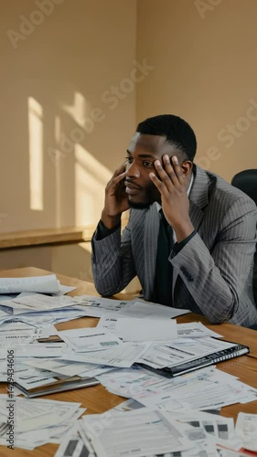 Worried business man with hands on head in front of messy desk covered in financial documents and paperwork, feeling stressed and overwhelmed.