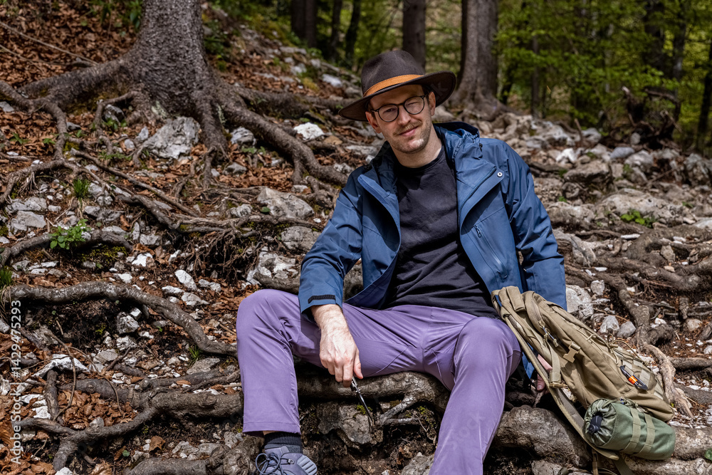 A young man of about thirty in a wide-brimmed hat sits on the roots of a tree in an alpine forest and is engaged in bushcrafting - whittling a wooden stick with a knife A young man of about thirty in a wide-brimmed hat sits on the roots of a tree in an alpine forest and is engaged in bushcrafting - whittling a wooden stick with a knife