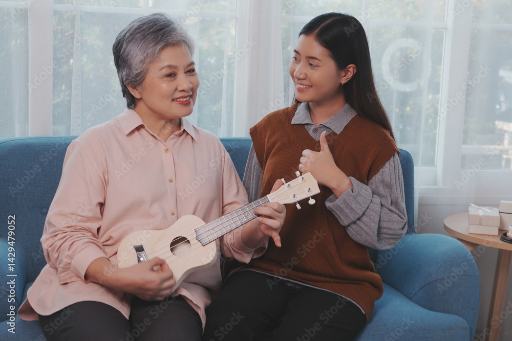Asian elderly mother and her beautiful 29-year-old daughter relax on the sofa playing ukulele happily together at home.