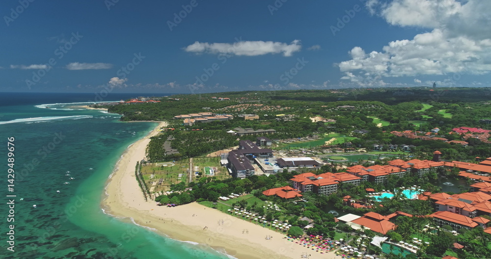 Stunning aerial drone footage captures the beauty of Nusa Dua beach with its luxury hotels, white sand, turquoise waters of the Indian Ocean, lush greenery, and a clear blue sky with white clouds